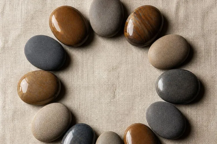 Twelve identical river pebbles arranged in a ring on linen, their finishes alternating between glossy and matte, illustrating the two alternating zodiac polarity groups