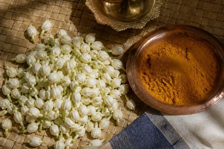 A brass oil lamp, loose jasmine buds, a copper bowl of turmeric and folded handloom cloth on a woven mat in morning light